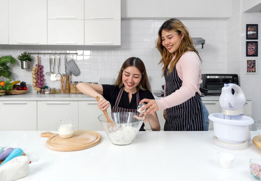 Two young women make dessert in the kitchen. One people stirring a white cake batter or icing mixture. The other pouring baking powder in a clear glass bowl