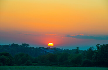Obraz premium Sunset over fields and trees in a green hilly landscape under a colorful sky in sunlight in summer, Voeren, Limburg, Belgium, June, 2024