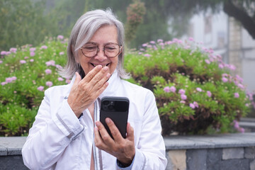 Video call. Grey haired laughing senior woman in garden outdoors using mobile phone video chatting. Old modern grandmother uses technology to stay in touch with family and friends