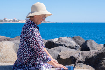 Senior woman with hat working on laptop sitting outdoor face to the sea - elderly nomadic caucasian female enjoying tech and social