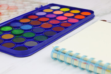 Water color for painting with blank sketch book and paint brush. White marble background with copy space.