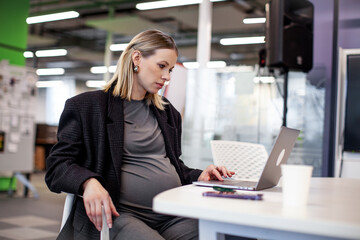 Confident pregnant female executive efficiently works on her computer in the office, seated at her...