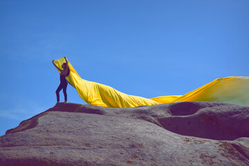 Freedom on the Peak. A girl stands atop a red rock, holding a yellow fabric that gracefully flutters in the wind. The scene embodies minimalism and the striking contrast of shapes and colors.