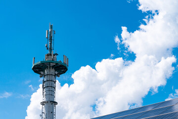 Broadcast mast and part of the roof with solar panels against a blue sky with white clouds.