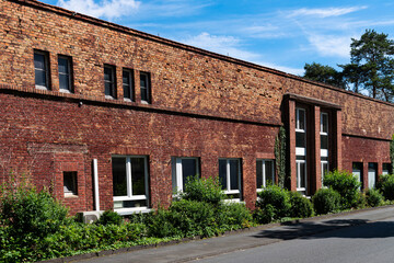 Wall of an old red brick industrial building with overgrown bushes under a blue sky with light white clouds.