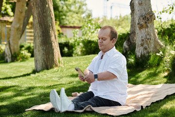 Fototapeta premium A man with inclusivity sits on a blanket in a park, using his phone.