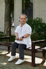Obraz premium A man with inclusivity sits on a bench, headphones on, enjoying a peaceful moment outdoors.