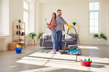 Happy young smiling married couple man and woman standing in the living room at home with vacuum...