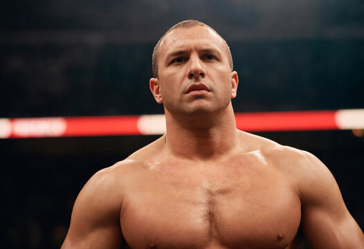 A muscular male boxer stands shirtless in a boxing ring, under the bright lights, with a determined look on his face.