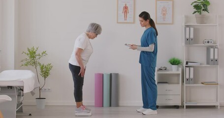 Female nurse or caregiver measuring weight on scales of elderly gray-haired woman in clinic and writing down results. Senior patient having consultation during exam in medical office. 4k video.
