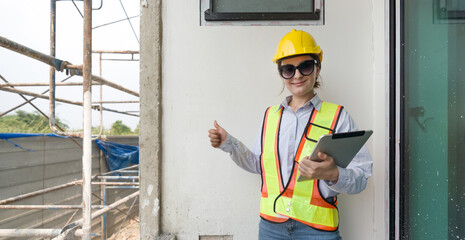 A construction worker is smiling, wearing safety gear, and holding a tablet, showing a thumbs-up at...