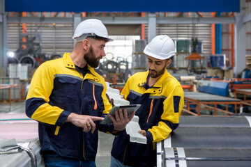 Two factory workers in work uniform and protective hardhat, inspecting a stack of large steel coils...