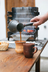 A hand pours hot coffee from a jezvah into a clay mug. A jezvah against the background of a brazier, in which fire and smoke are visible.