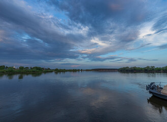 A small boat cruises along a calm river at sunset, with a backdrop of vibrant, spring foliage and a sky full of dramatic, wispy clouds.