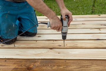 A carpenter kneels on a newly constructed wooden platform, using a cordless drill to screw a fastener into the planks.