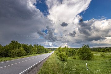 A winding road cuts through a lush forest, disappearing into the distance under a dramatic, cloudy sky. The light from the sun shines through the clouds, casting long shadows over the trees and road.