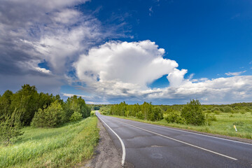 A scenic road winds through a field of green grass and trees with a blue sky and fluffy white clouds overhead.