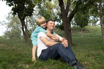 Fototapeta premium Portrait of a happy dad and son. A cute boy 6-7 years old hugs his father sitting on the grass. Walk in the park. Father's Day, Son's Day. Tender relationships in the family