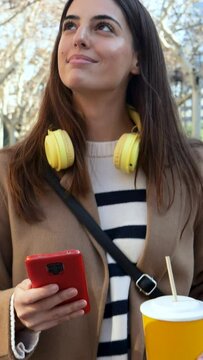 Vertical video. Elegant smiling young woman walking through a city with a fruit juice in her hands and a mobile phone