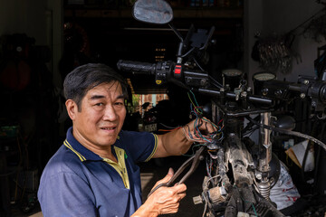 Vietnamese man looking at the camera while working in his motorcycle workshop repairing the electric circuit. Half of his body is in the shade