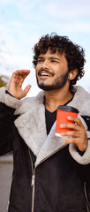 Young indian man in earphones holding coffee outdoors, vertical banner