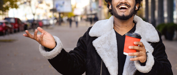 Cheerful indian man in earphones holding coffee in city, banner