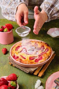 Sprinking of a sugar powder of a round pie from the dough of the Philo with strawberries and cream in pink form for baking on a green concrete background.