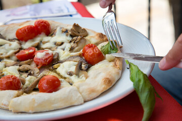 A person is using a fork to slice a pizza into pieces. Round pizza marinara with cherry tomato, mozzarella, mushrooms and anchovy. carbohydratesraditional italian food. Homemade food. Carboidrate 