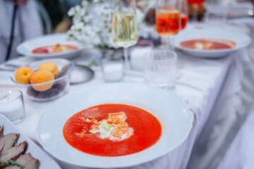 A close-up of a gazpacho plate and a covered festive table with a white tablecloth. Delicious tomato soup, glasses of wine and fruits.