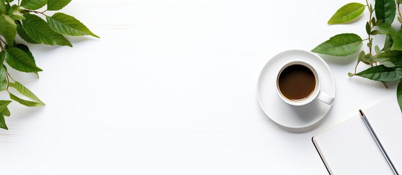 An office desk viewed from above with items like a notebook, book, supplies, pencil, leaf, and coffee cup arranged neatly on the white surface, forming a visually appealing copy space image.