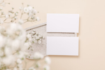 Business paper cards on ceramic tray with gentle gypsophila flowers on beige background, top view. Mockup paper card for design, print, wedding and invitation.