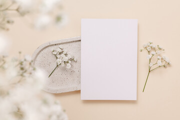 Festive paper greeting card on ceramic tray and gentle gypsophila flowers on beige background, top view. Mockup paper card for design, print, wedding and invitation