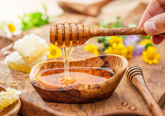 Honey dripping from wooden spoon into wooden bowl with honey, spring flowers are on wooden table...