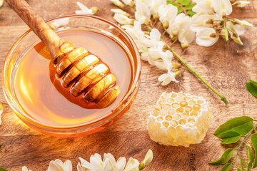 Dipping wooden spoon into glass bowl with honey, acacia flowers are on wooden table near. Top view.