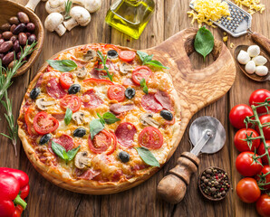 Pizza topped with cherry tomatoes, salami and other ingredients is on wooden table surrounded by ingredients. Top view.