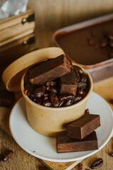 Cocoa bean tea packet in a white glass on a wooden table with chocolate chunks next to it surrounded by seeds.