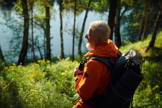 High angle view of senior male tourist wearing orange jacket with gray backpack looking at forest lake, copy space