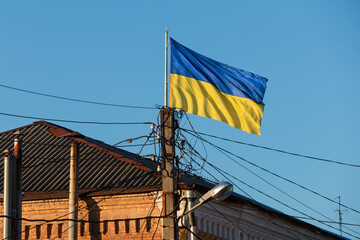 Flag of Ukraine and a concrete pole with overhead electricity wires near an old building. Ukrainian flag flutters in the wind