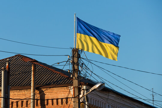 Flag of Ukraine and a concrete pole with overhead electricity wires near an old building. Ukrainian flag flutters in the wind