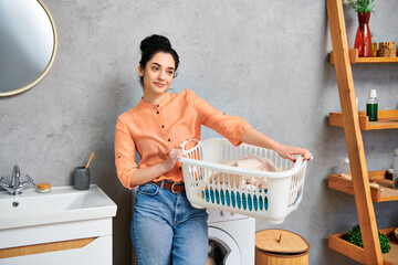 A stylish woman in casual attire holding a laundry basket standing next to a washer, preparing to...