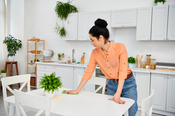 A stylish woman in casual attire meticulously wipes down a table in a home kitchen, creating a...