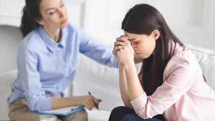 Fototapeta premium A young woman sits with her head in her hands, visibly distressed, while a therapist observes and offers comfort in a serene, private counseling room.