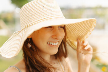 A woman wearing a hat and with long hair is happily smiling in a park, looking relaxed and carefree on a sunny day