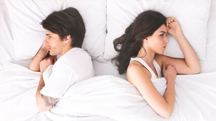 A young couple lies in bed, turned away from each other, seemingly after an argument. They are both lying on their sides, with their backs to each other