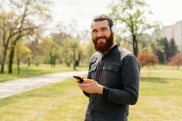 Fototapeta premium Smiling bearded man using smartphone outdoors in a park on a sunny day, showcasing technology and lifestyle.