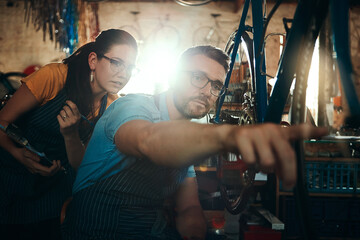 Bicycle technician, working and pointing in shop with frame, chain and tools for maintenance or cycling. Man, bike and mechanic in workshop as teamwork for gear, inspection and safety with glasses