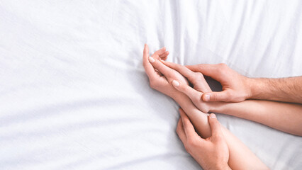This image shows the hands of a couple intertwined on a white bed, suggesting intimacy and connection, cropped, copy space
