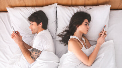 A man and a woman are lying in bed, each using their smartphones. The couple is lying on their sides, facing away from each other. The image is taken from above, looking down on the couple.