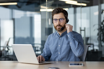 Obraz premium Portrait of a serious young man in a headset and glasses sitting at a desk in the office, holding a microphone with his hand, working on a laptop, looking confidently into the camera