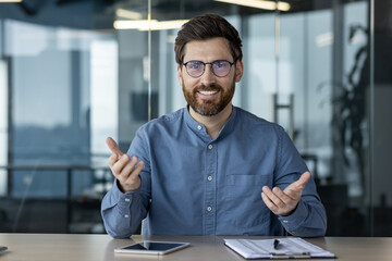 Obraz premium Online conference and business meeting in the office. A young smiling man in glasses sits in front of the camera and talks on a video call, gesturing with his hands
